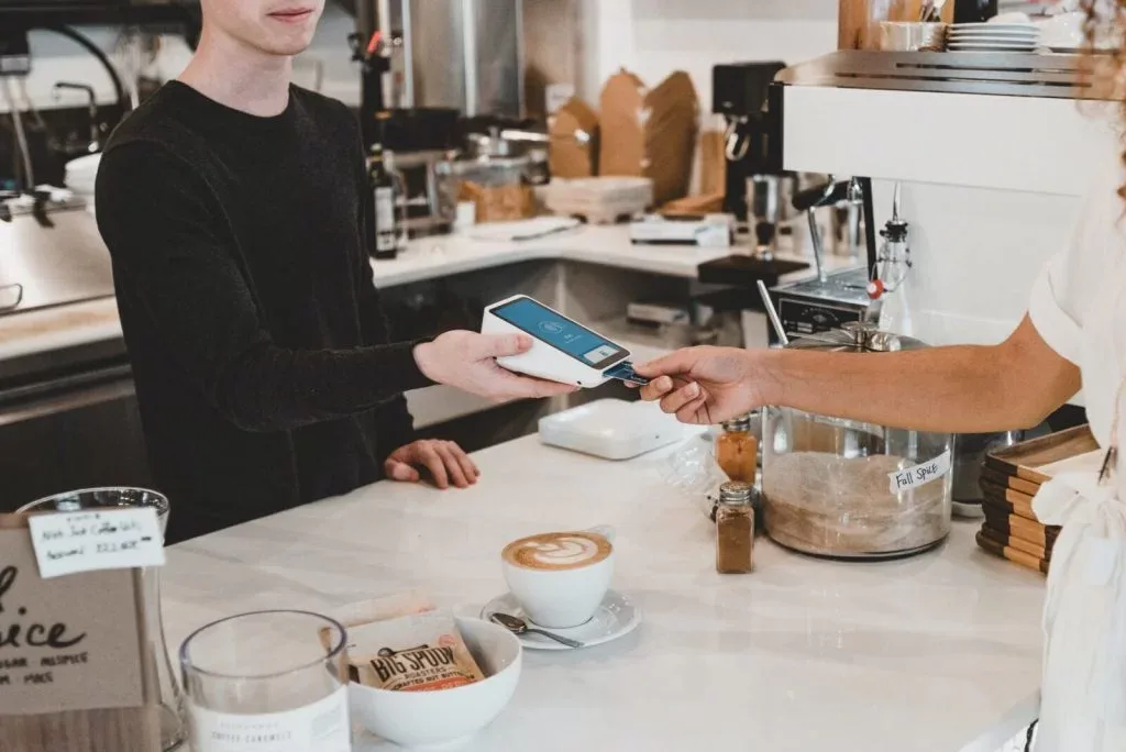 A barista handing a card terminal across a café counter, a latte waiting beside it.