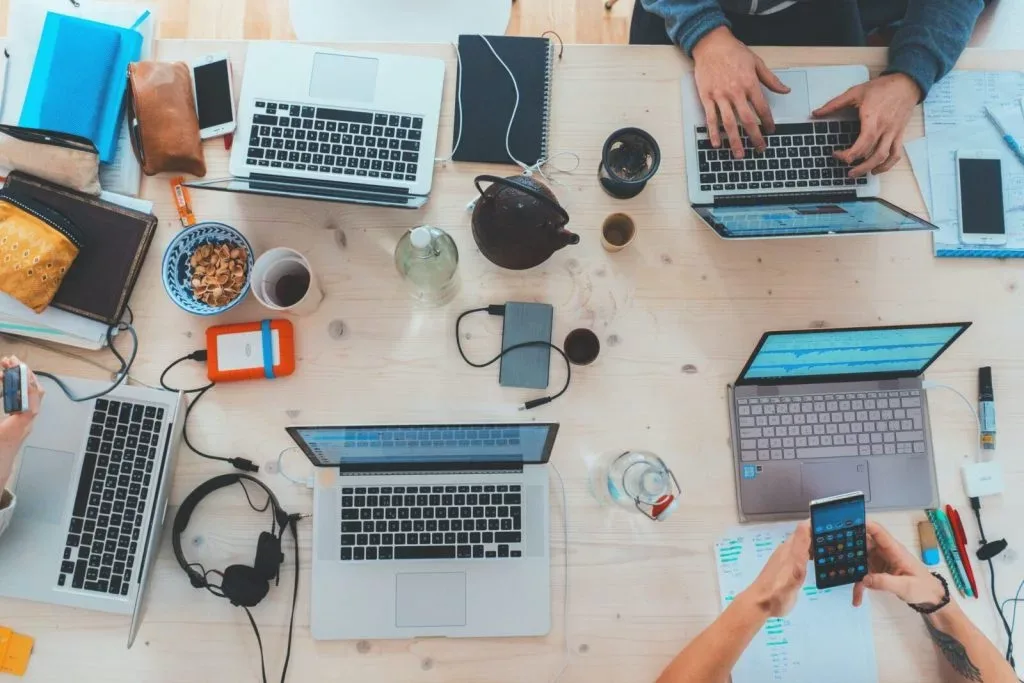 Wspólne biurko widziane z góry: cztery otwarte laptopy, notatniki, słuchawki i czajniczek.