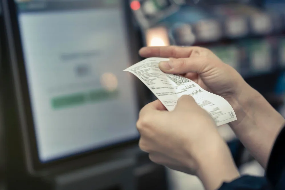 Hands holding a paper till receipt in front of a payment terminal at a shop counter.