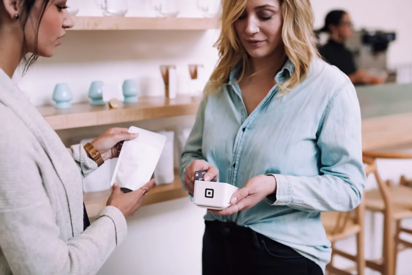 A customer holding a card reader and a bank card at a café counter while an assistant hands over a paper pouch.