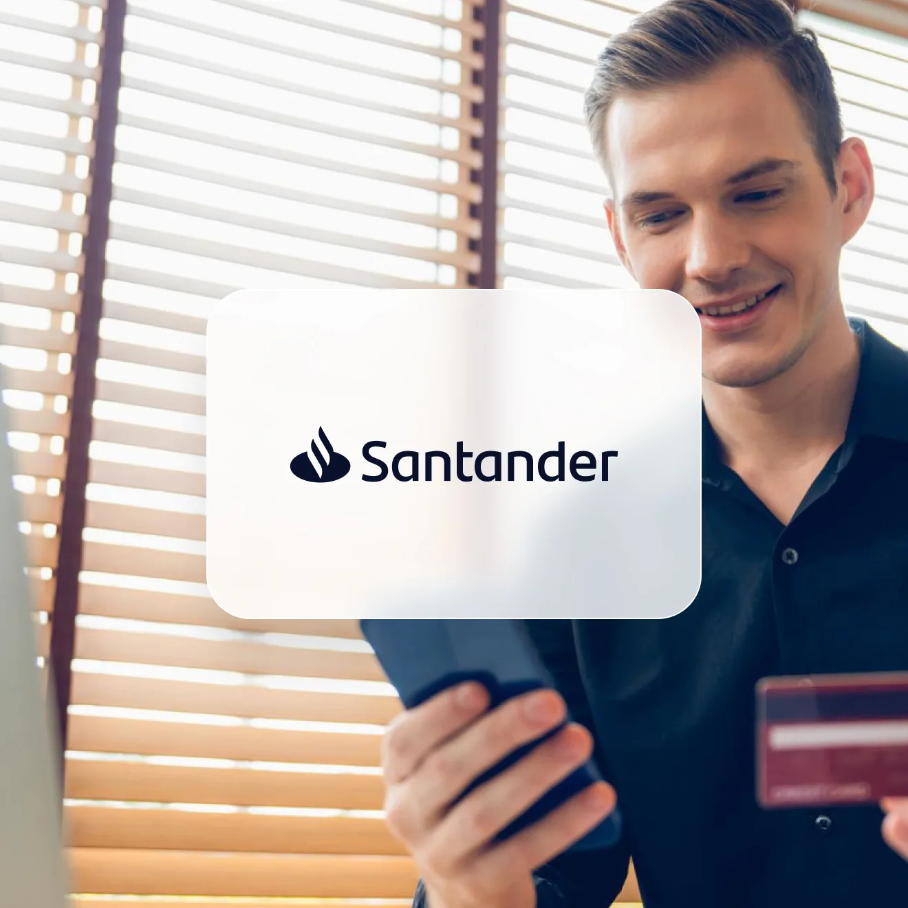 Smiling man in a dark shirt looking at his phone and holding a payment card, in a room with wooden blinds, with the Santander logo on a card over the photo.