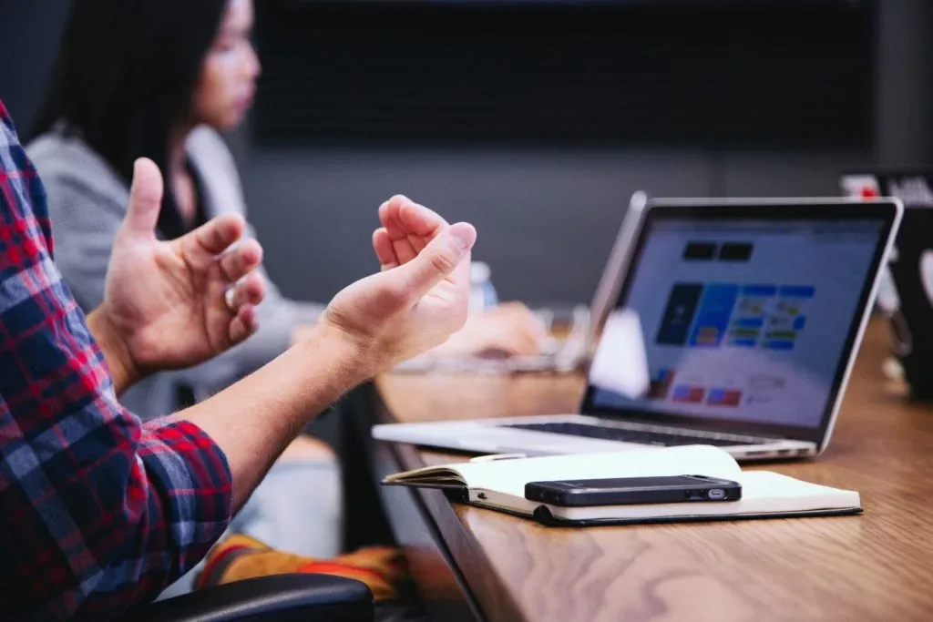 Someone gesturing with both hands while talking across a meeting table, an open laptop showing a dashboard beside them.
