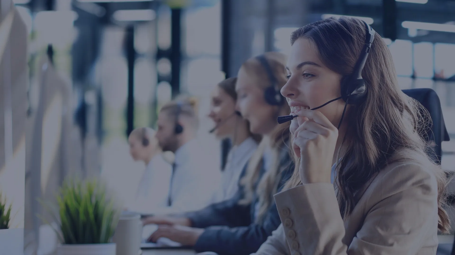 A row of contact-centre agents in headsets at their desks, the one nearest the camera smiling as she speaks