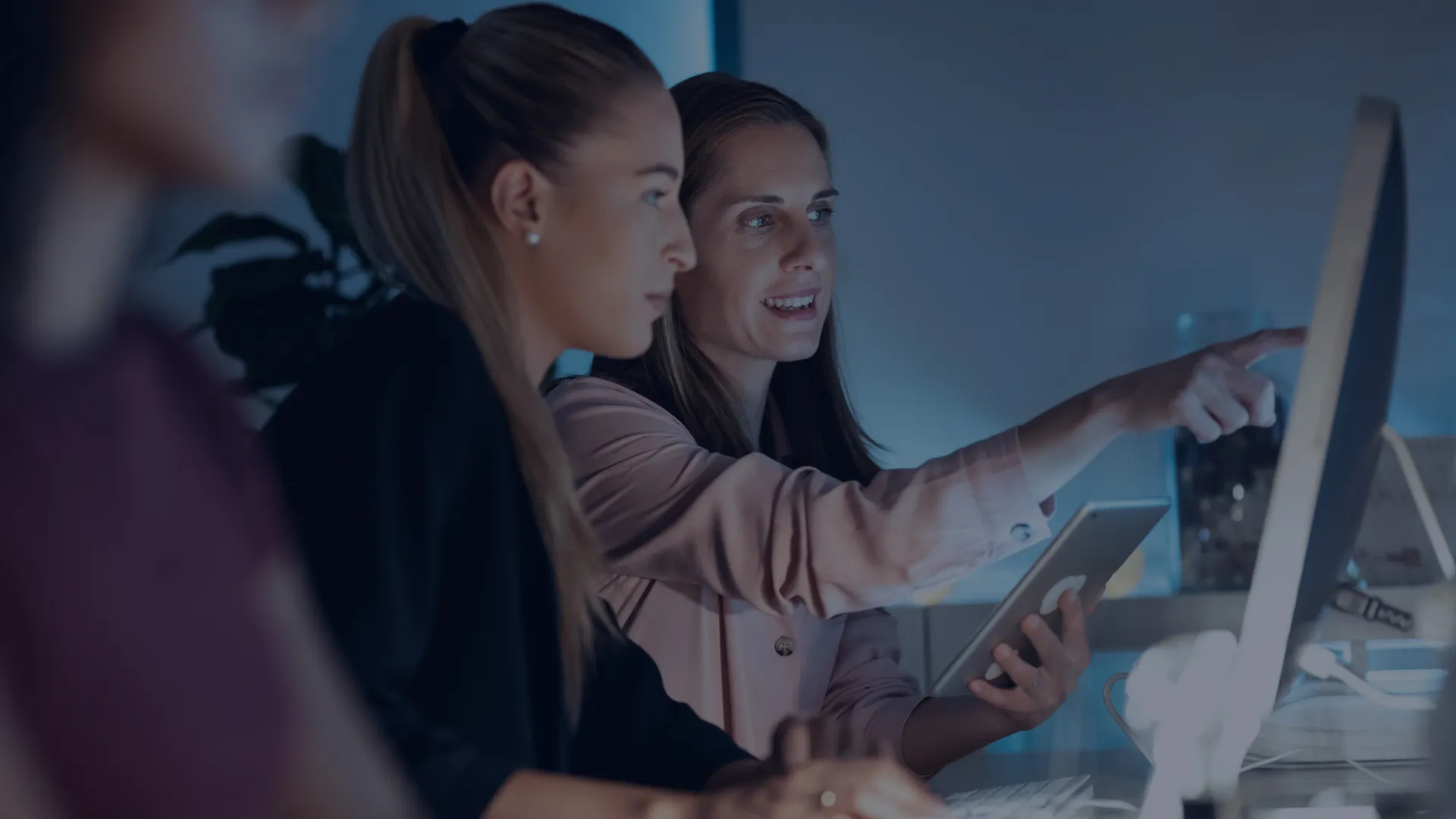 Two colleagues at a desk in a dimly lit office looking at a monitor, one of them pointing at the screen
